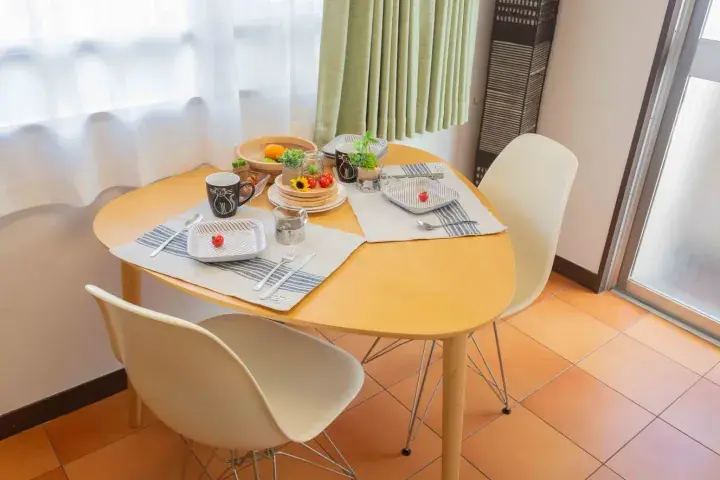 A bright dining area with a round wooden table set for two, featuring white chairs and natural light from a window.