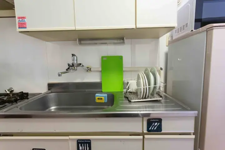 Compact kitchen sink area with a green cutting board, dish rack, and white cabinets in a modern apartment.