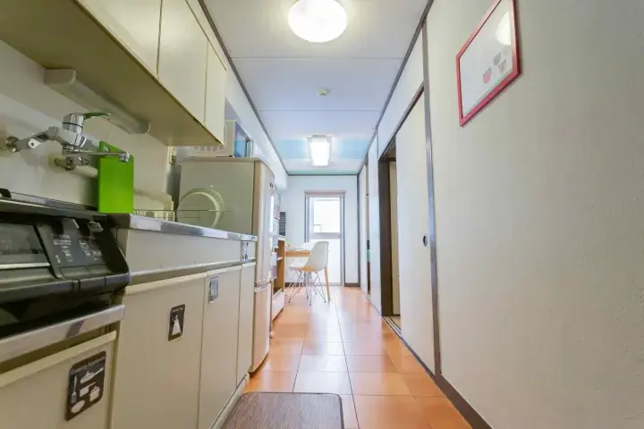 Narrow kitchen corridor with white cabinets, appliances, and tiled floor leading to a dining area.