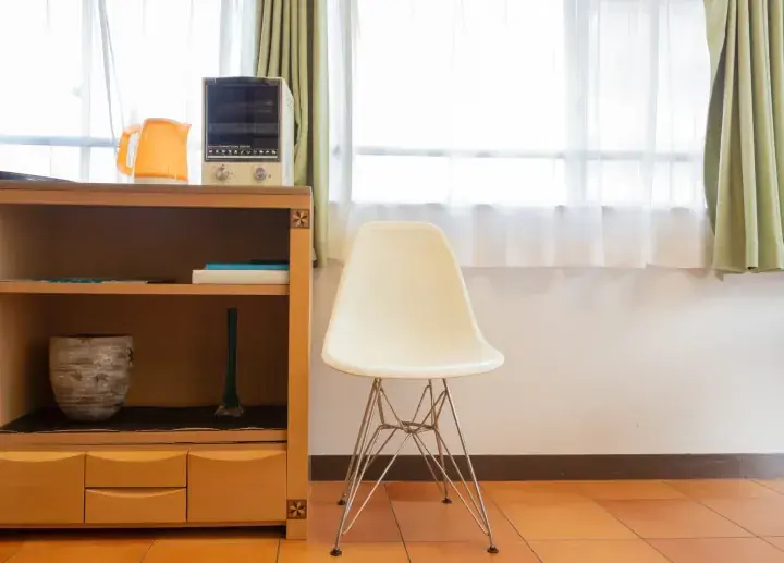 White molded plastic chair with wire base beside wooden cabinet in bright room with window and curtains