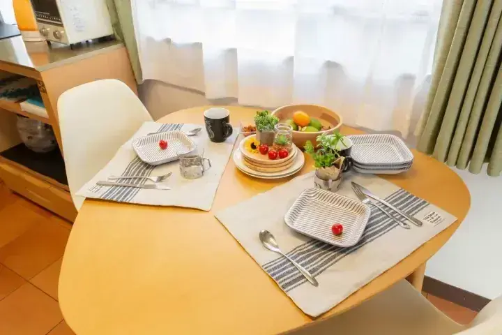 白いカーテンの窓際に置かれた、プレートとカトラリー、果物が並んだモダンなダイニングテーブル。