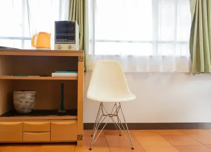 窓とカーテンのある明るい部屋に置かれた、木製キャビネットの横にある白いモールドプラスチックチェア