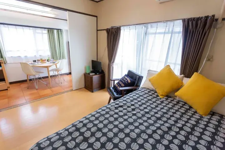 Bright bedroom with patterned bedspread and yellow pillows, overlooking a dining area with a round table and chairs.