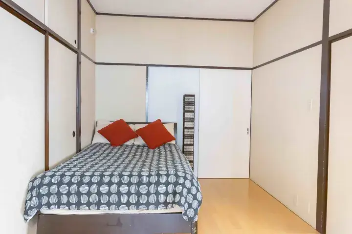 A bedroom with a bed featuring a patterned duvet and orange pillows, set against white walls and wooden flooring.