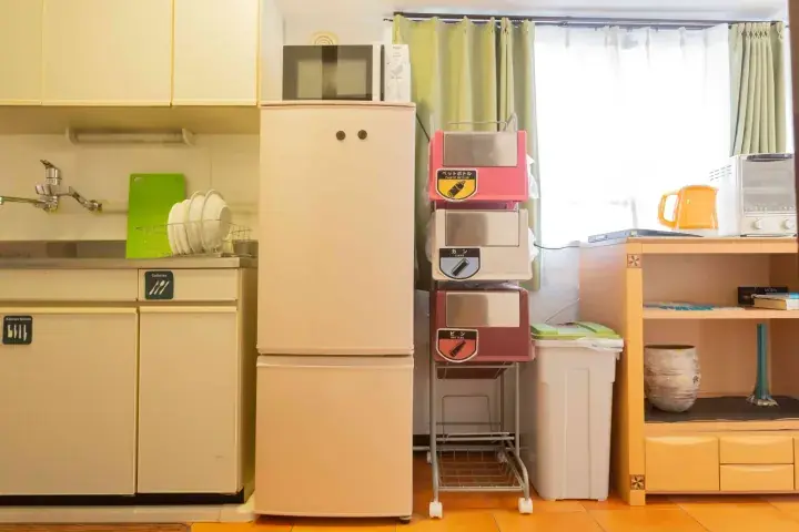 Compact kitchen interior featuring a cream refrigerator, microwave, dish rack, and colorful storage units near a window.