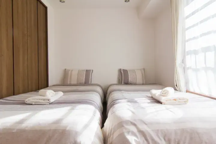 Bright twin bedroom with neatly made beds, striped bedding, and fresh towels near a window with sheer curtains.