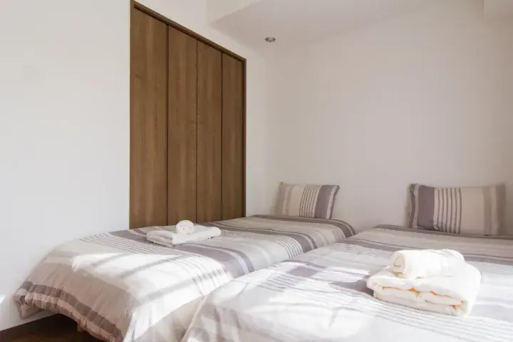 Bright bedroom with two single beds, striped bedding, and a wooden wardrobe in a minimalist Malaysian property.