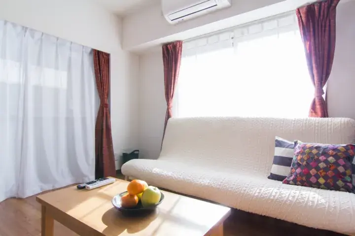 Bright living room with a white sofa, wooden coffee table with fruit, and large windows with sheer and patterned curtains.