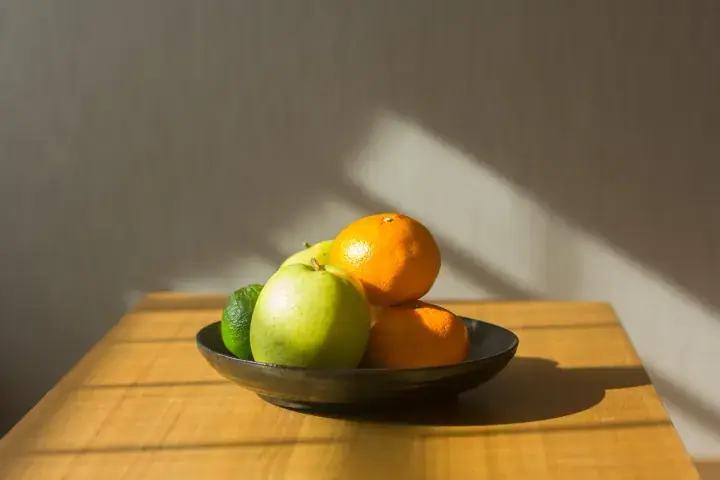 A black bowl of fresh fruit including oranges and apples sits on a wooden table illuminated by warm sunlight.