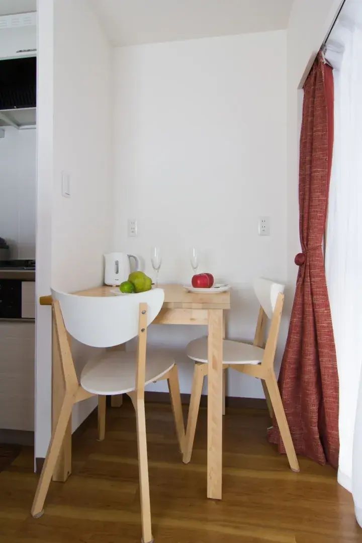 Minimalist dining area with a light wood table, white chairs, and fruit, featuring a red curtain and kitchen access.
