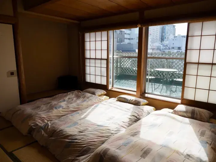 Traditional Japanese tatami room with futons, shoji screens, and a balcony view of city buildings.