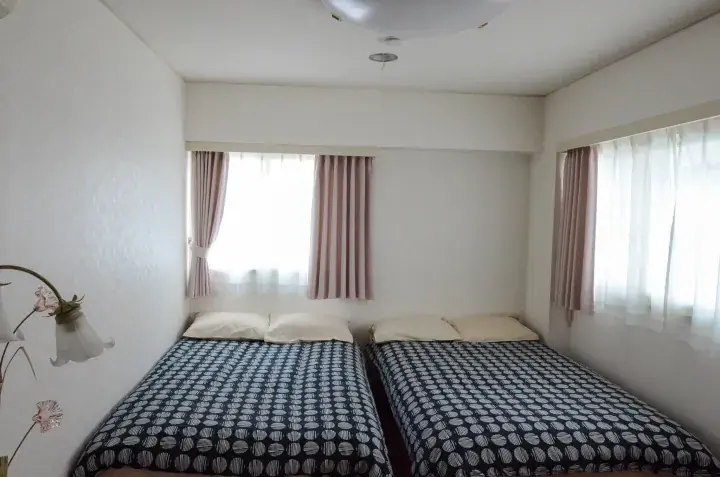 Bright twin bedroom with patterned beds, pink curtains, and natural light from two windows.