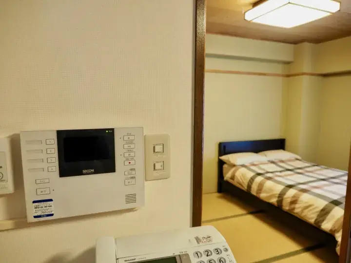 Interior view of a Japanese-style room with a SECOM security panel, telephone, and a bed with a plaid blanket.