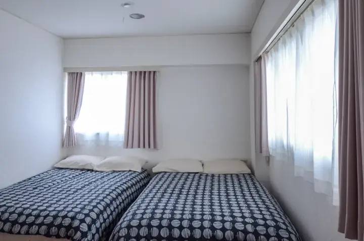 Bright twin bedroom with patterned bedding and large windows draped in sheer and solid curtains.