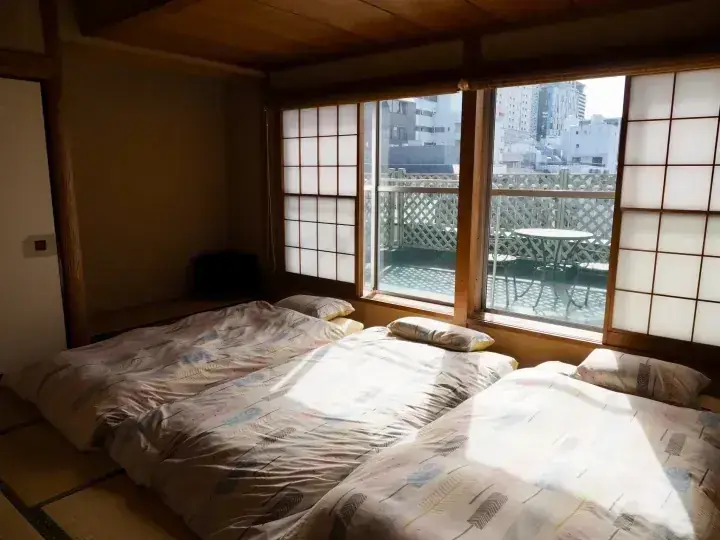 畳の和室で布団が敷かれ、障子と窓から都市の景色が見える。