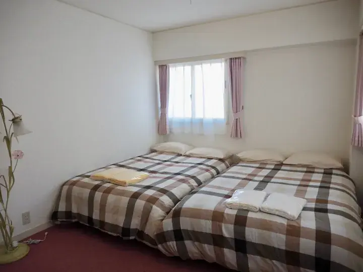 Two single beds with plaid bedding in a bright hotel room with pink curtains and a floor lamp