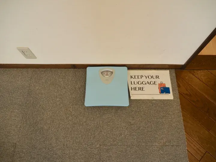 A light blue bathroom scale sits on a carpeted floor next to a sign reading KEEP YOUR LUGGAGE HERE.