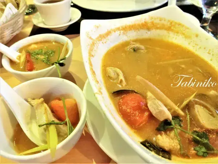 A large bowl and two smaller bowls of Tom Yum soup with chicken, tomatoes, and herbs, served at a Malaysian restaurant.