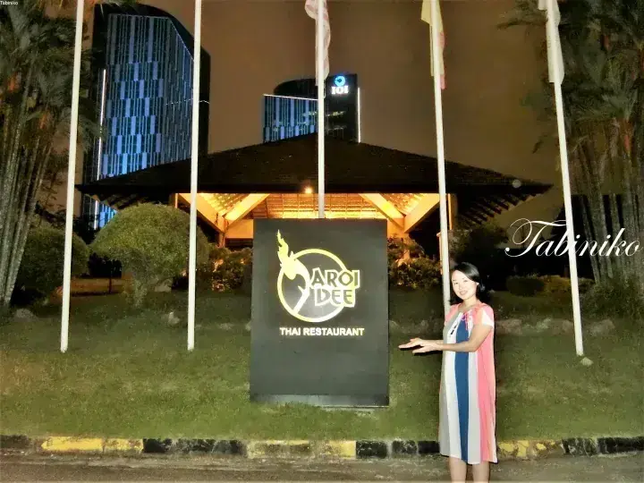 A woman poses beside the illuminated Aroi Dee Thai Restaurant sign at night, with IOI City Mall towers visible in the background.