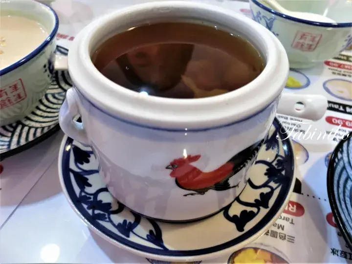 A white ceramic cup with a rooster design holds dark herbal tea, served on a matching saucer at a Malaysian restaurant.