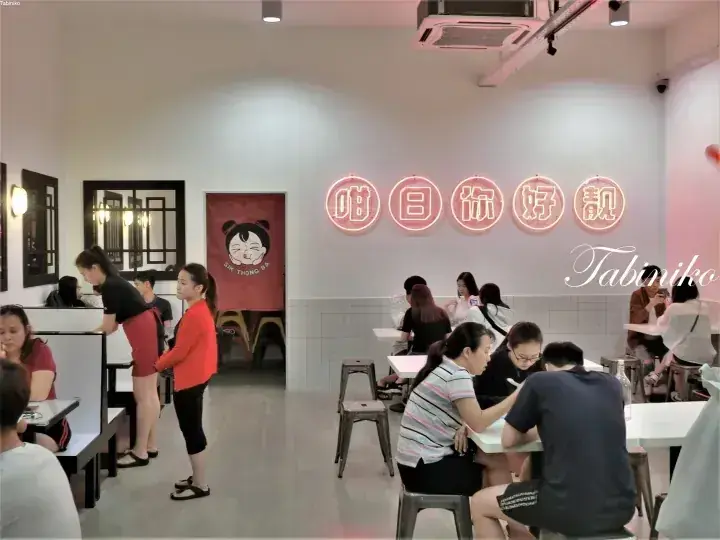 壁に「Gai Thong」と書かれたピンクのネオンサインとキャラクターロゴがある店内で食事をする客たち。