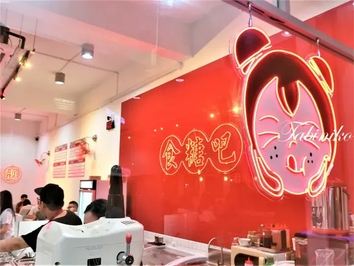 Bright red wall with neon sign featuring a cartoon character and Chinese characters at a busy food stall counter.