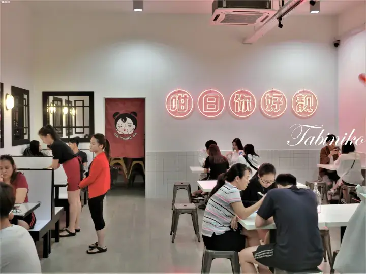 Customers dining inside a restaurant with a pink neon sign reading “Gai Thong” and a cartoon logo on the wall.