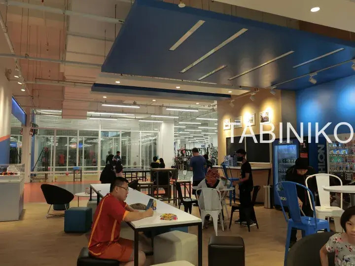 Customers relaxing at a modern gym cafe area with blue ceiling panels and glass-walled workout zones.