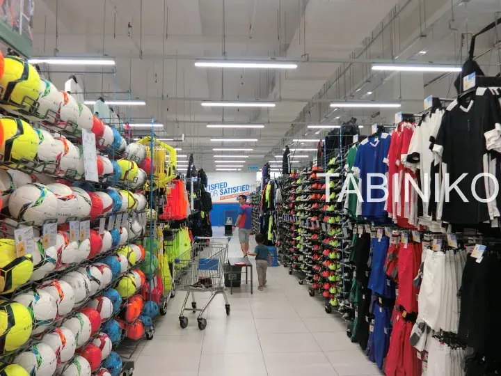 Aisle in a sports store filled with colorful soccer balls and jerseys, with customers browsing.