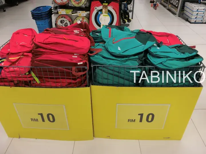 Red and green backpacks displayed in yellow bins with RM 10 price tags at a Malaysian retail store.
