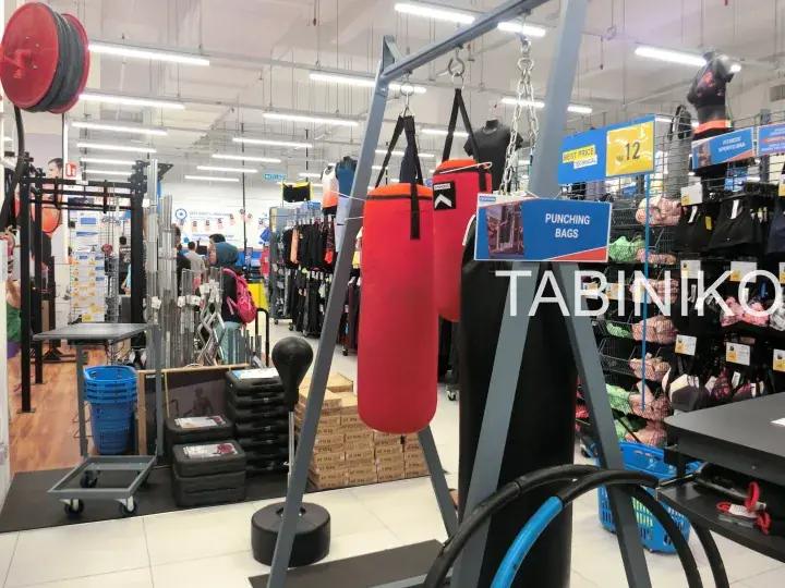 Red punching bags hanging from a metal stand in a sports retail store aisle with exercise equipment nearby