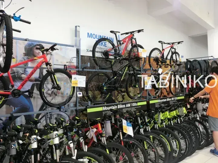 デカトロン店内のラックに展示されたマウンテンバイクと、AM 100sの看板、そして買い物客の姿。