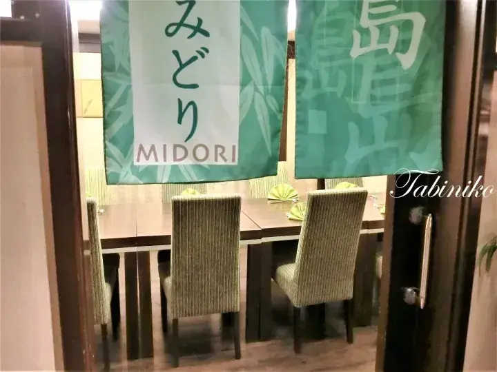Restaurant interior with green noren curtains reading MIDORI and 島, wooden tables, and striped chairs.