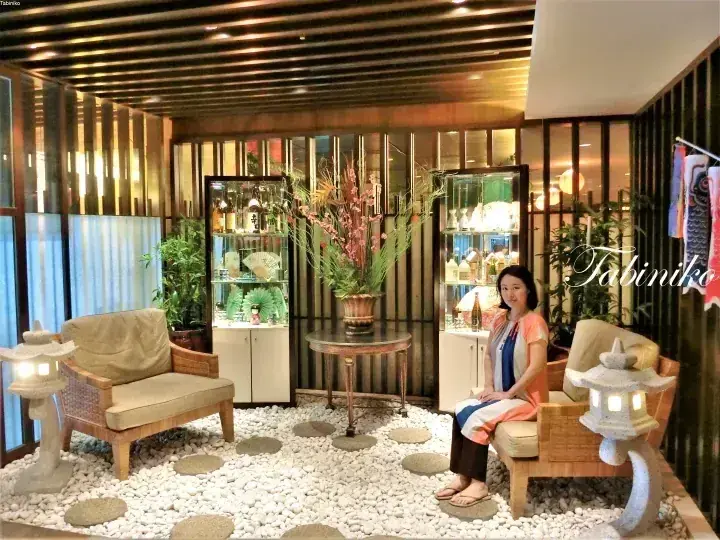 Woman sitting on a wooden chair in a Japanese-style lounge with white pebbles, stone lanterns, and floral arrangements.