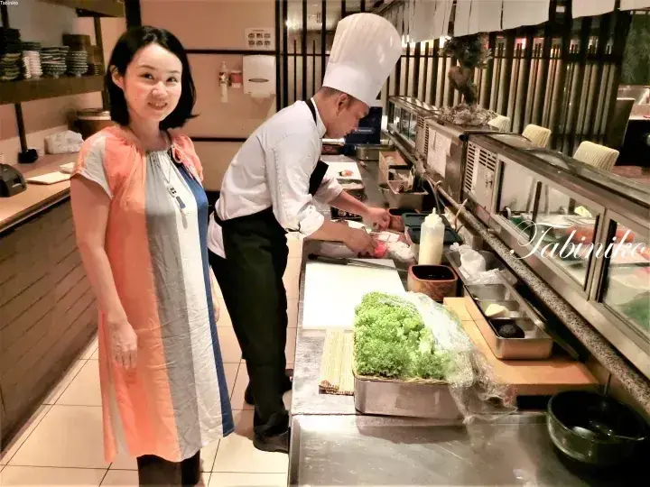 A woman stands smiling beside a sushi chef preparing food at the counter in a Japanese restaurant.