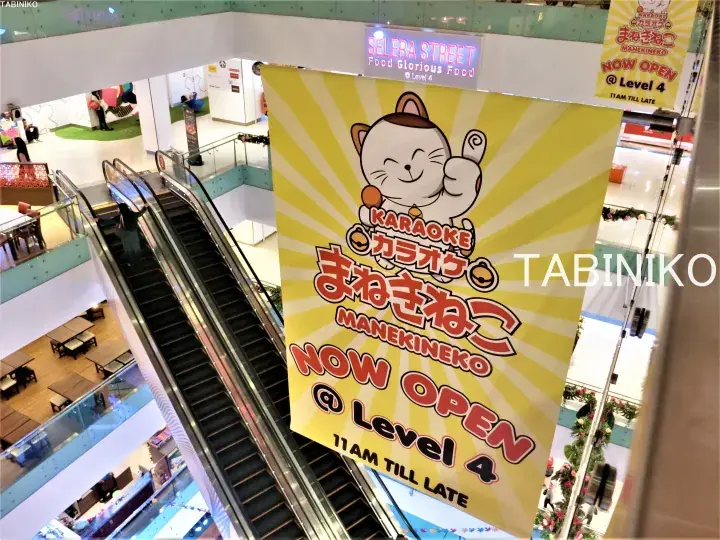 Yellow banner for Manekineko Karaoke hanging in a mall atrium near escalators.