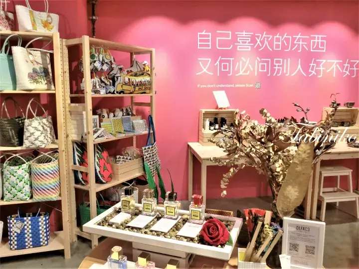 A boutique shop interior with pink walls, wooden shelves displaying woven bags, and a perfume display featuring a red rose.
