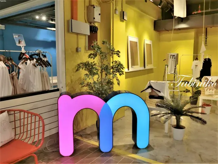 Illuminated pink and blue &rsquo;m&rsquo; sign in a bright yellow clothing store with racks of white garments and potted plants.