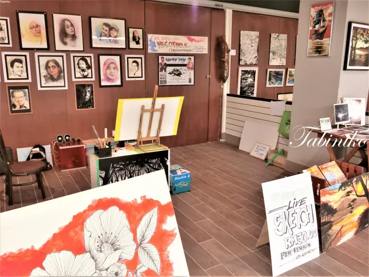 Interior of Tabinka art studio with framed portraits on walls, an easel with canvas, and a sign advertising live sketching services.
