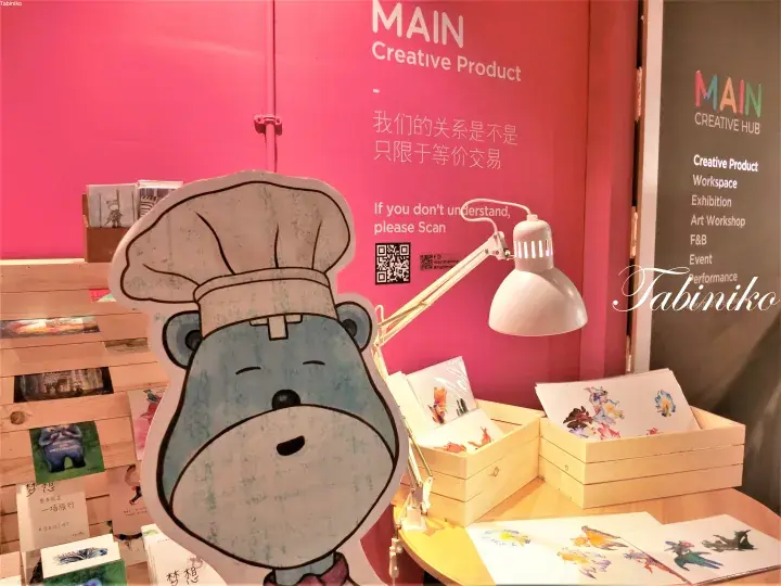 A cartoon bear mascot wearing a chef hat stands beside a display of MAIN Creative Product items and a pink wall with text.