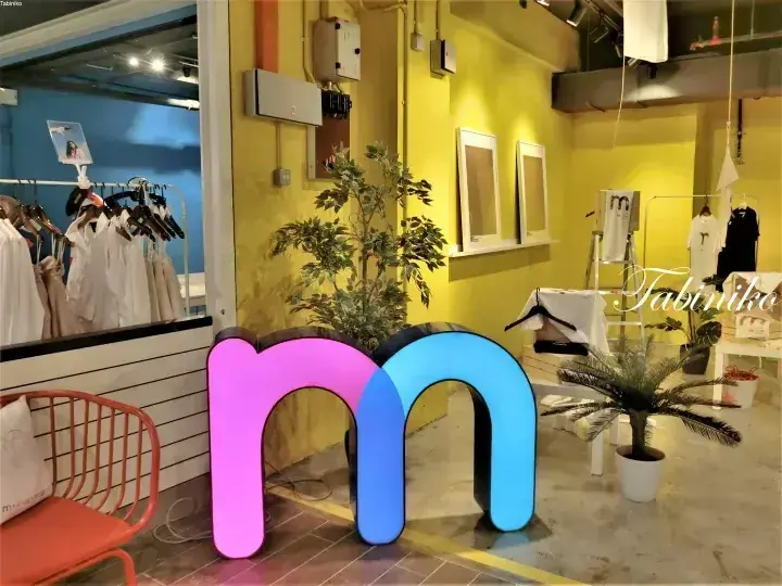 白い衣類や観葉植物が並ぶ、黄色い壁の衣料品店内に設置されたピンクと青の発光する「m」のサイン。