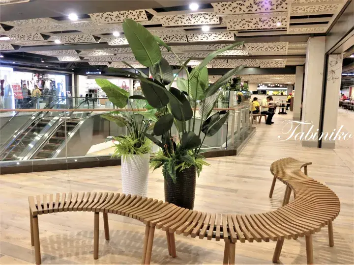 Curved wooden bench and potted plants in a modern shopping mall atrium with decorative ceiling and escalators.