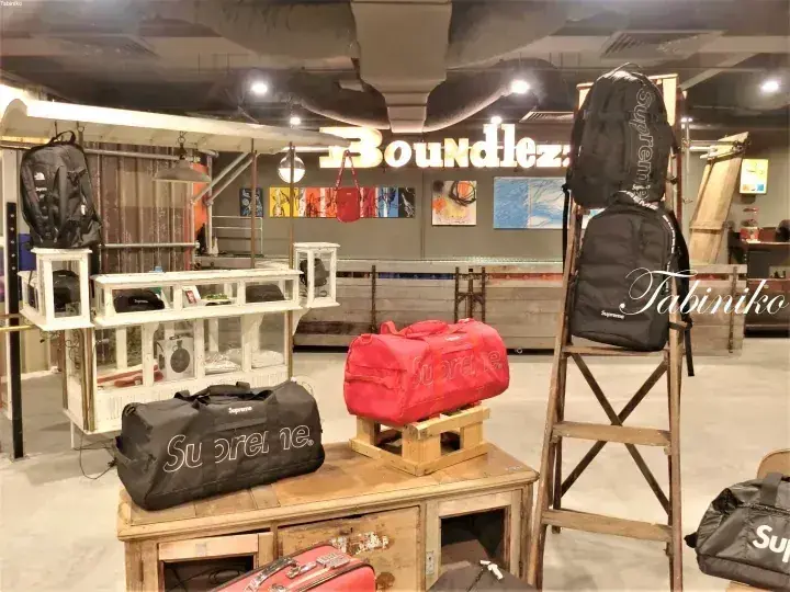 照明サインが見えるBundlez店内に木製パレットやはしごに展示されたSupremeのバッグ類。