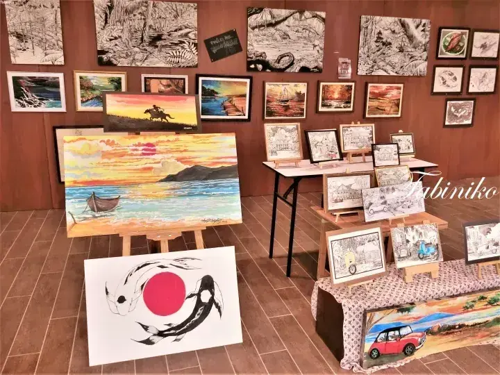 イーゼルやテーブルに展示された、色鮮やかな風景画、白黒スケッチ、そして大きな日本の国旗アートが並ぶアート展示会。