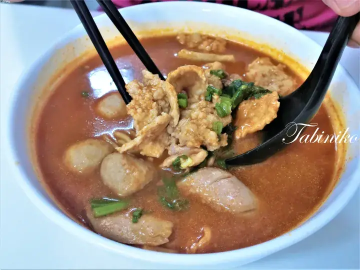 A bowl of spicy laksa with fish balls, pork slices, and greens being lifted by chopsticks and a spoon.