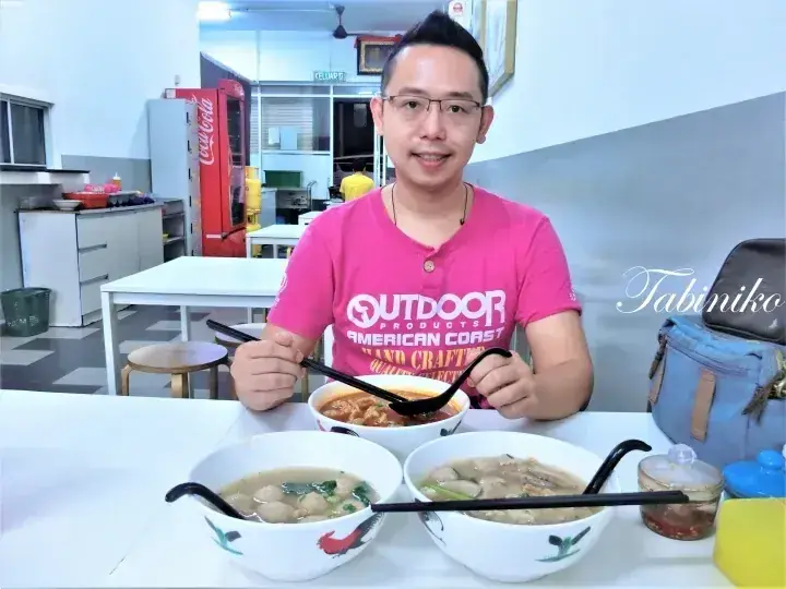 カジュアルな食堂で、ピンクのシャツを着た男性が麺料理の前で笑顔を見せる。