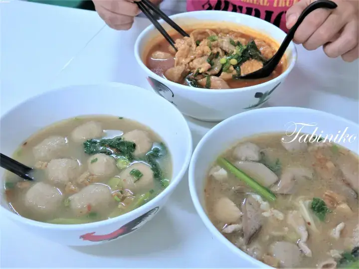 Three bowls of Malaysian noodle soup with meatballs and greens on a white table, featuring a red curry bowl being eaten with chopsticks.