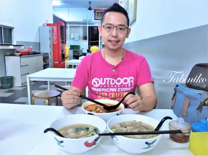 A man in a pink shirt smiles while holding a spoon over bowls of noodles at a casual eatery.