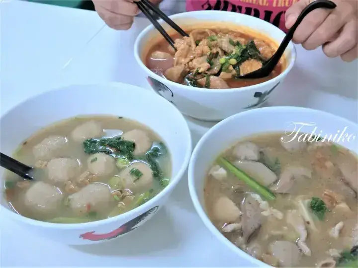 白いテーブルに並べられた、肉団子と野菜が入ったマレーシアの麺料理3皿。箸で赤いカレー味の麺を食べている様子。
