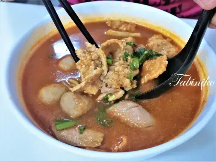 箸とスプーンで魚肉団子や豚肉をすくう、具だくさんのスパイシーなラーサのボウル。